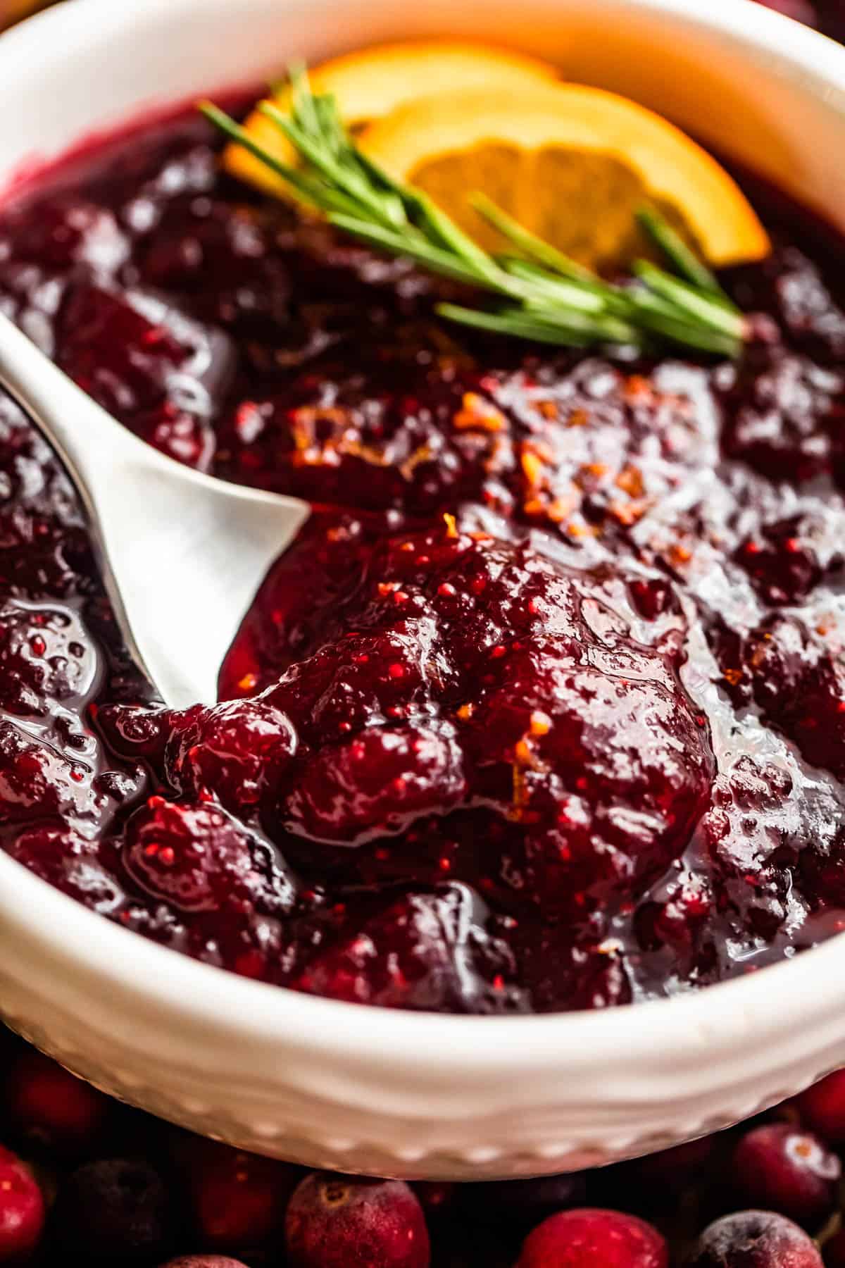 side shot of a spoon inside of a bowl filled with cranberry sauce and garnished with rosemary sprig plus orange slices