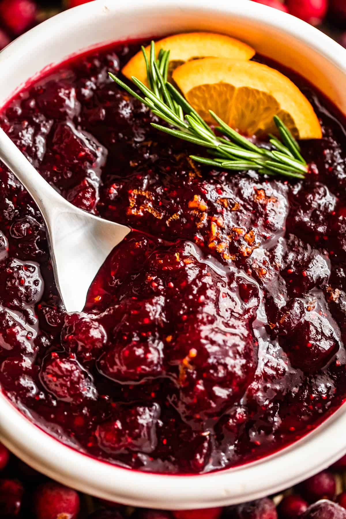 overhead close up shot of a spoon inside of a bowl filled with cranberry sauce and garnished with rosemary sprig plus orange slices