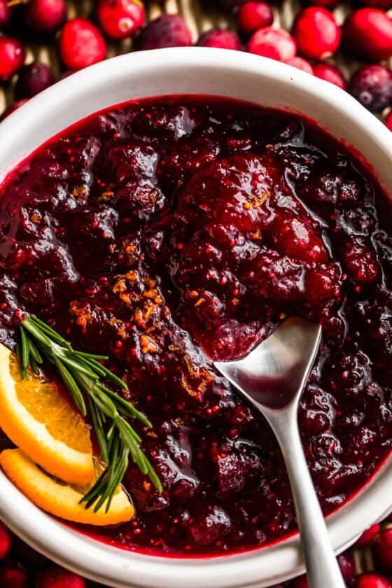 overhead close up shot of a spoon inside of a bowl filled with cranberry sauce and garnished with rosemary sprig plus orange slices