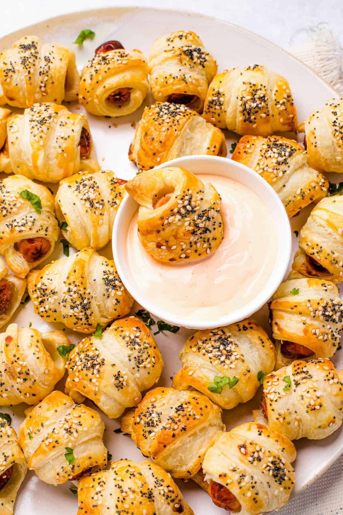 A platter of pigs in a blanket with a bowl of dipping sauce in the center. One pastry is dipped in the sauce.