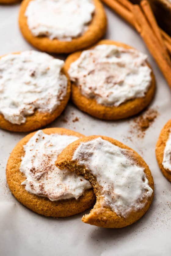 Frosted Pumpkin Cookies arranged on a white background with one cookie halfway bitten off and a few cinnamon sticks set at the top of the right corner.