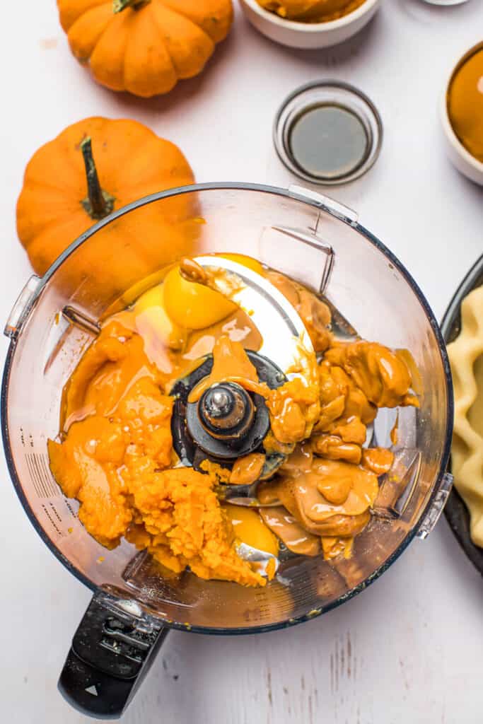 Pumpkin filling ingredients in a blender on a white work surface, with mini pumpkins decorating the surface.