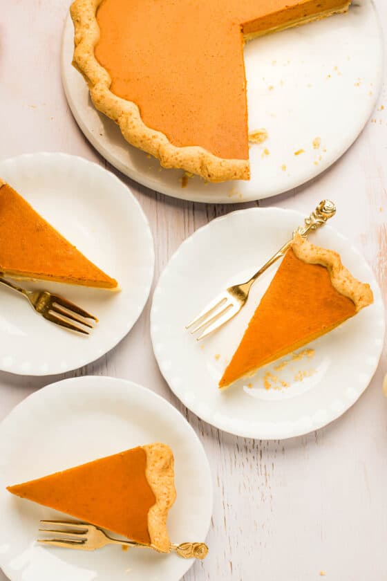 A pumpkin pie in a white serving platter. One slice has been cut and placed on a dessert plate with a golden fork.