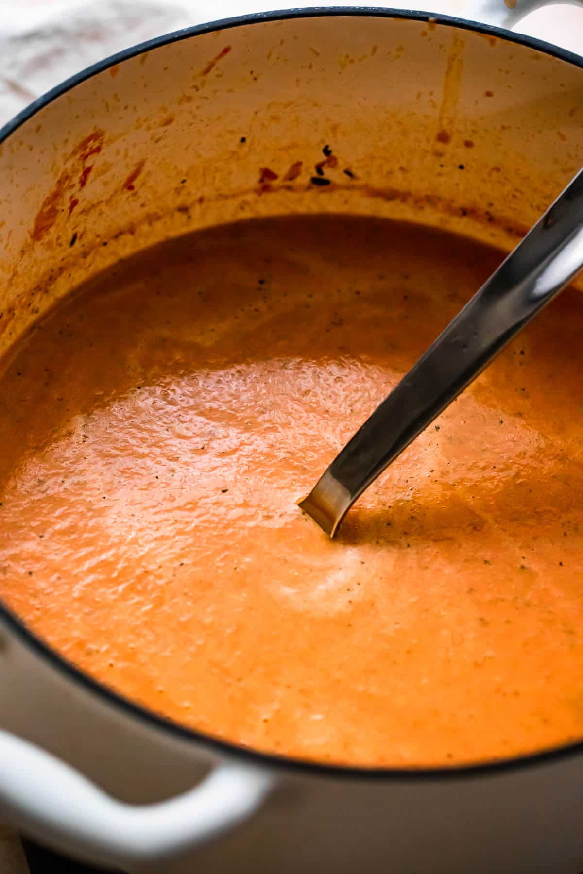 side shot of a white dutch oven with tomato basil soup and a ladle inside the soup.