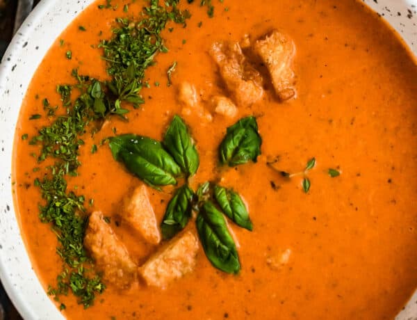 overhead shot of white bowl with tomato basil soup, garnished with croutons, parsley, and basil leaves.