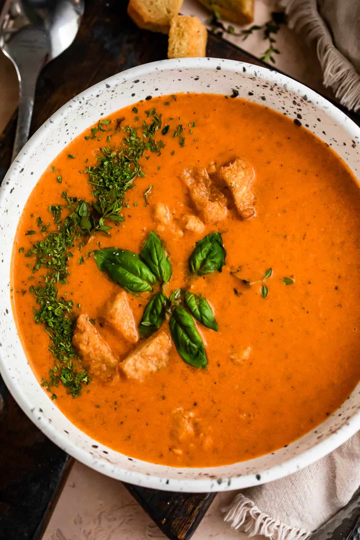 overhead shot of white bowl with tomato basil soup, garnished with croutons, parsley, and basil leaves.