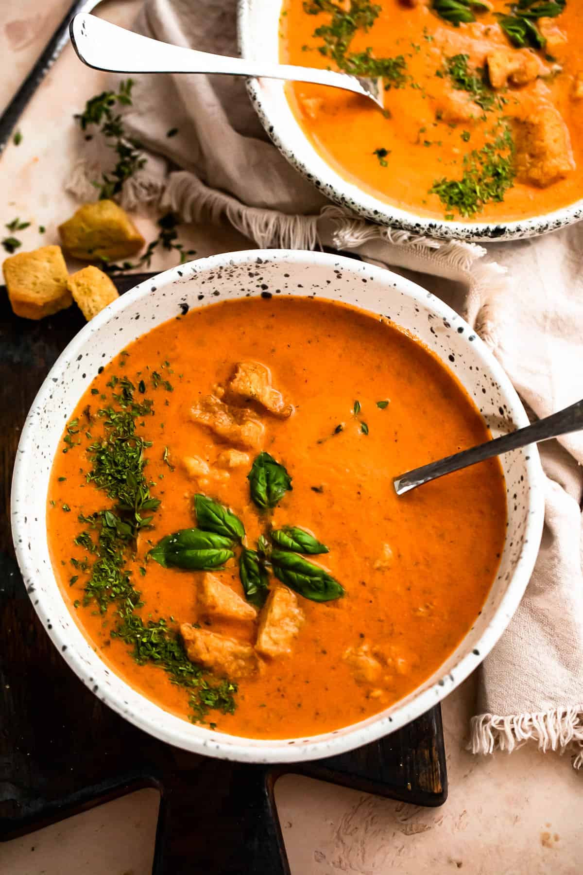 overhead shot of two white bowl with tomato basil soup, garnished with croutons, parsley, and basil leaves.