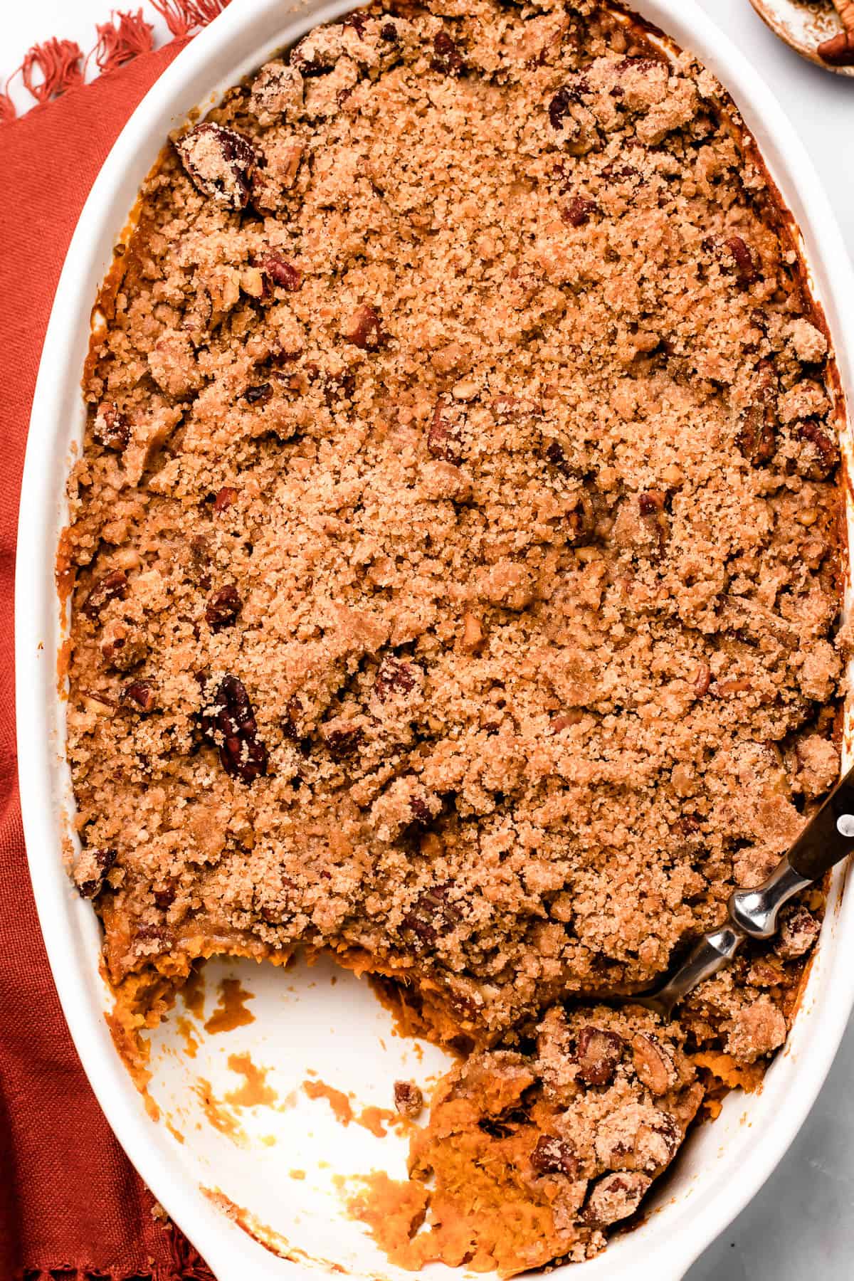 A baking dish of sweet potatoes and crumb topping, on a marble background. A serving of the casserole has been scooped out, and a spoon is resting in the dish.