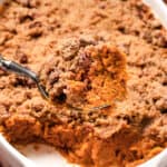 A shot of the sweet potato casserole with a serving spoon lifting a portion out of the dish, showing the texture of the baked casserole mixture and crumb topping.