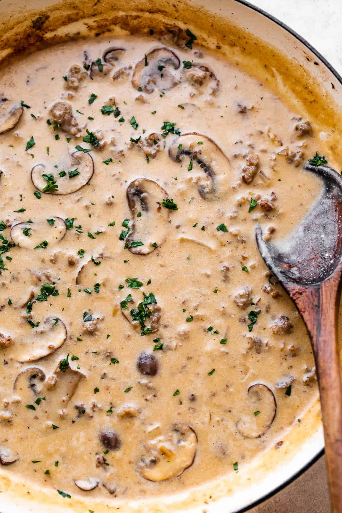 white skillet with simmering beef and mushrooms in a cream sauce with a wooden spoon inside the skillet.