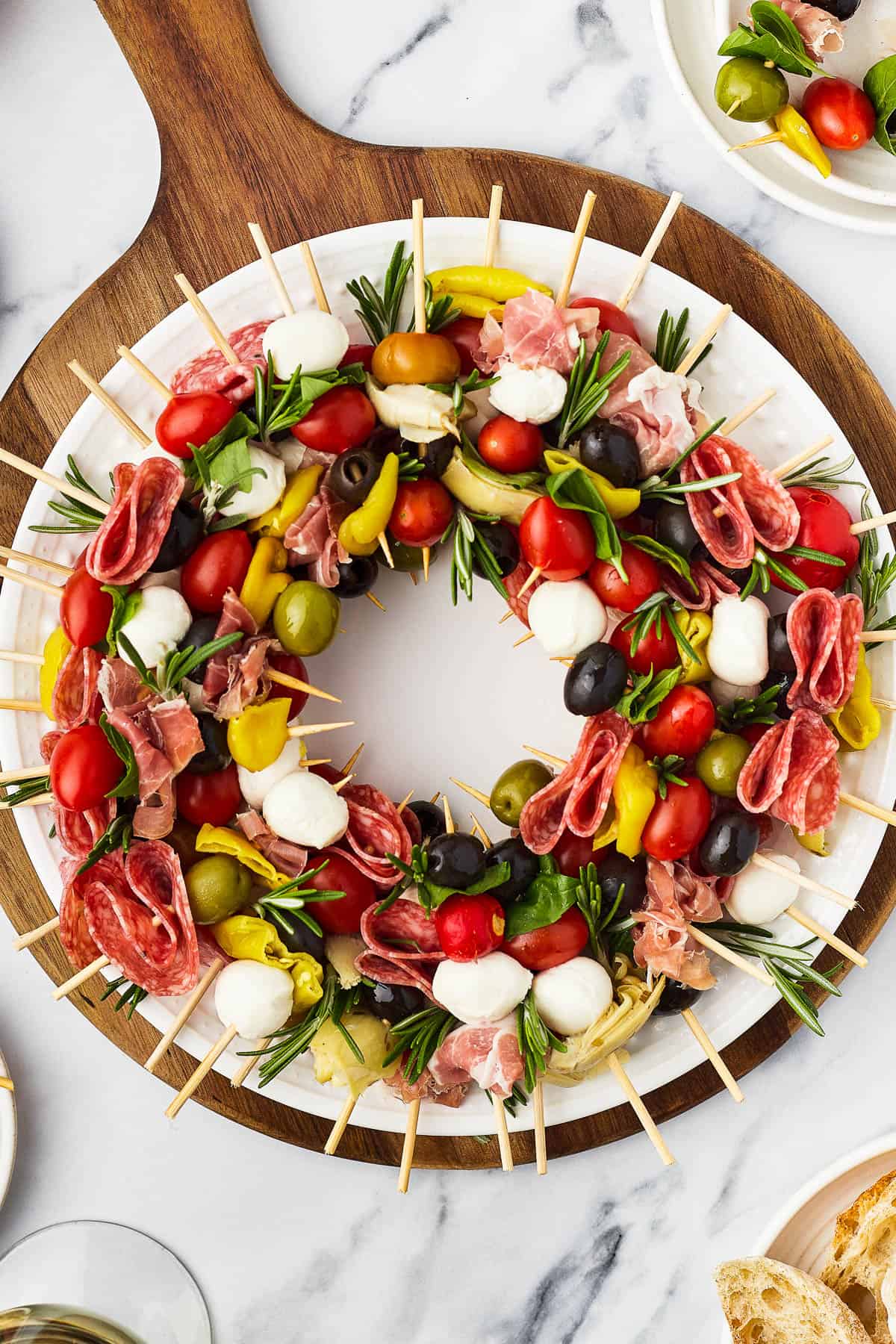 A table with a cutting board holding a wreath of skewers, a small pair of scissors, and fresh rosemary sprigs.