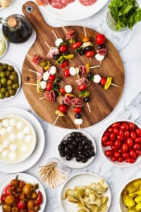Skewers of olives, mozzarella, etc. on a round cutting board, surrounded by bowls of antipasto ingredients.