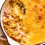 A spoon lifting a serving of creamed corn pudding from a white cast iron skillet.