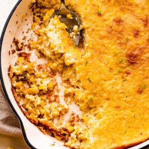 A spoon lifting a serving of creamed corn pudding from a white cast iron skillet.