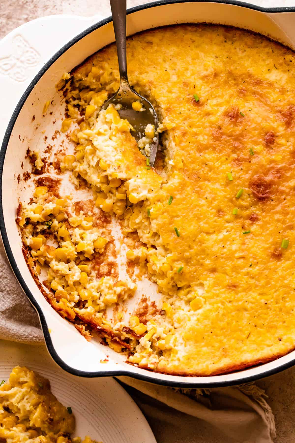 A spoon lifting a serving of creamed corn pudding from a white cast iron skillet.