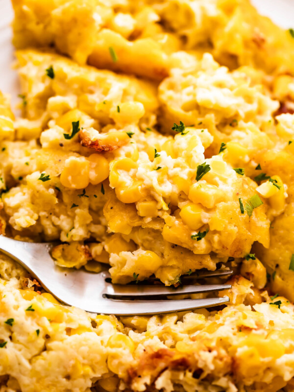 A shallow dish holding a serving of creamed corn casserole, near a baking dish of the same casserole. A spoon is in the bowl as well.