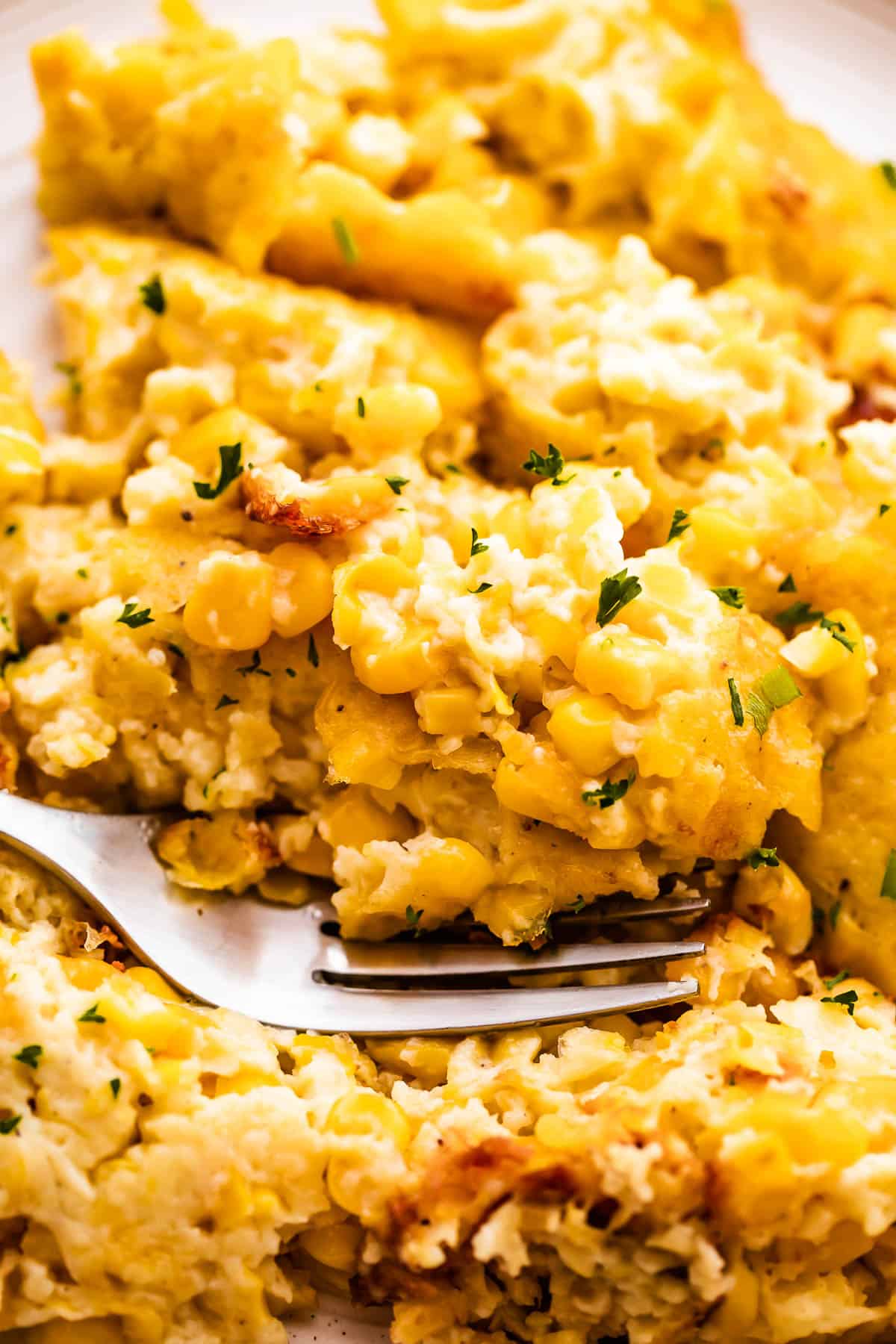 A shallow dish holding a serving of creamed corn casserole, near a baking dish of the same casserole. A spoon is in the bowl as well.