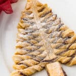 A baked puff pastry Christmas tree, dusted with powdered sugar, on an oval platter. Christmas greenery, a red bow, and a small bowl of chocolate dipping sauce surround the platter.