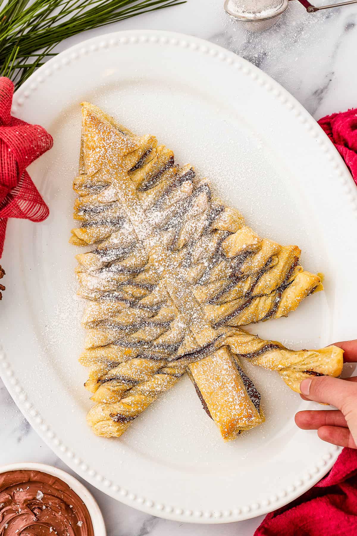 A baked puff pastry Christmas tree, dusted with powdered sugar, on an oval platter. Christmas greenery, a red bow, and a small bowl of chocolate dipping sauce surround the platter.