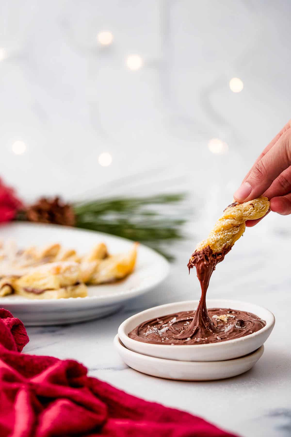 A woman's hand dips a puff pastry twist into chocolate dipping sauce.Christmas decorations and a platter are in the background of this shot.