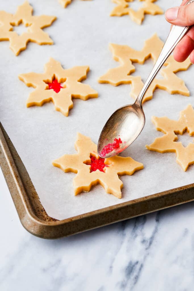 Cut out snowflake cookies with stars cut out of the center. A spoon is filling the centers with crushed red candy.