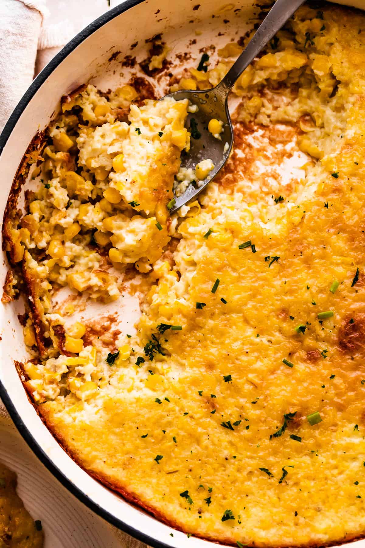 A spoon lifting a serving of creamed corn pudding from a white cast iron skillet.