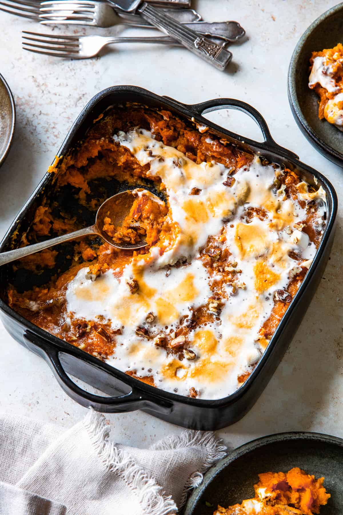 a square black baking dish filled with sweet potato casserole, topped with melted marshmallows. The marshmallow topping is golden-brown from baking. Several portions have been scooped out, and a serving spoon rests in the empty part of the dish.