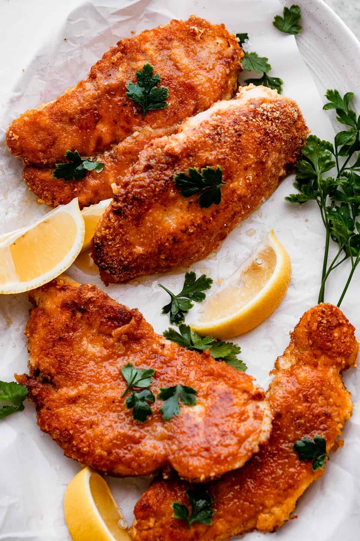 four slices of chicken schnitzel arranged on a white oval plate with lemon wedges set around the chicken and a garnish of parsley arranged over the chicken breast schnitzel.