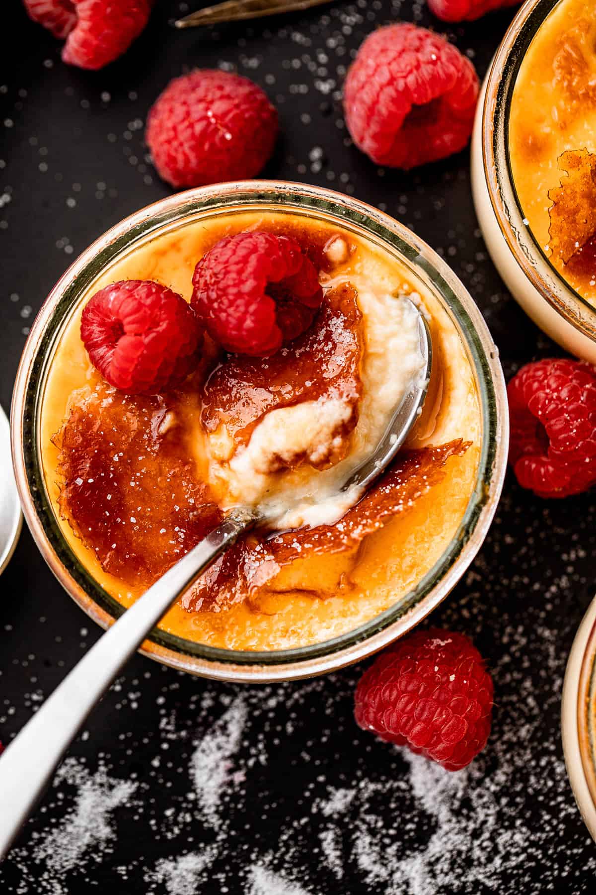 overhead shot of creme brulee in a glass jar, topped with two raspberries, and a spoon inside the jar.