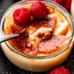 side shot of creme brulee in a glass jar, topped with two raspberries, and a spoon inside the jar.