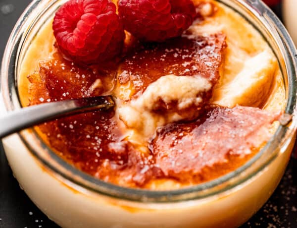 side shot of creme brulee in a glass jar, topped with two raspberries, and a spoon inside the jar.