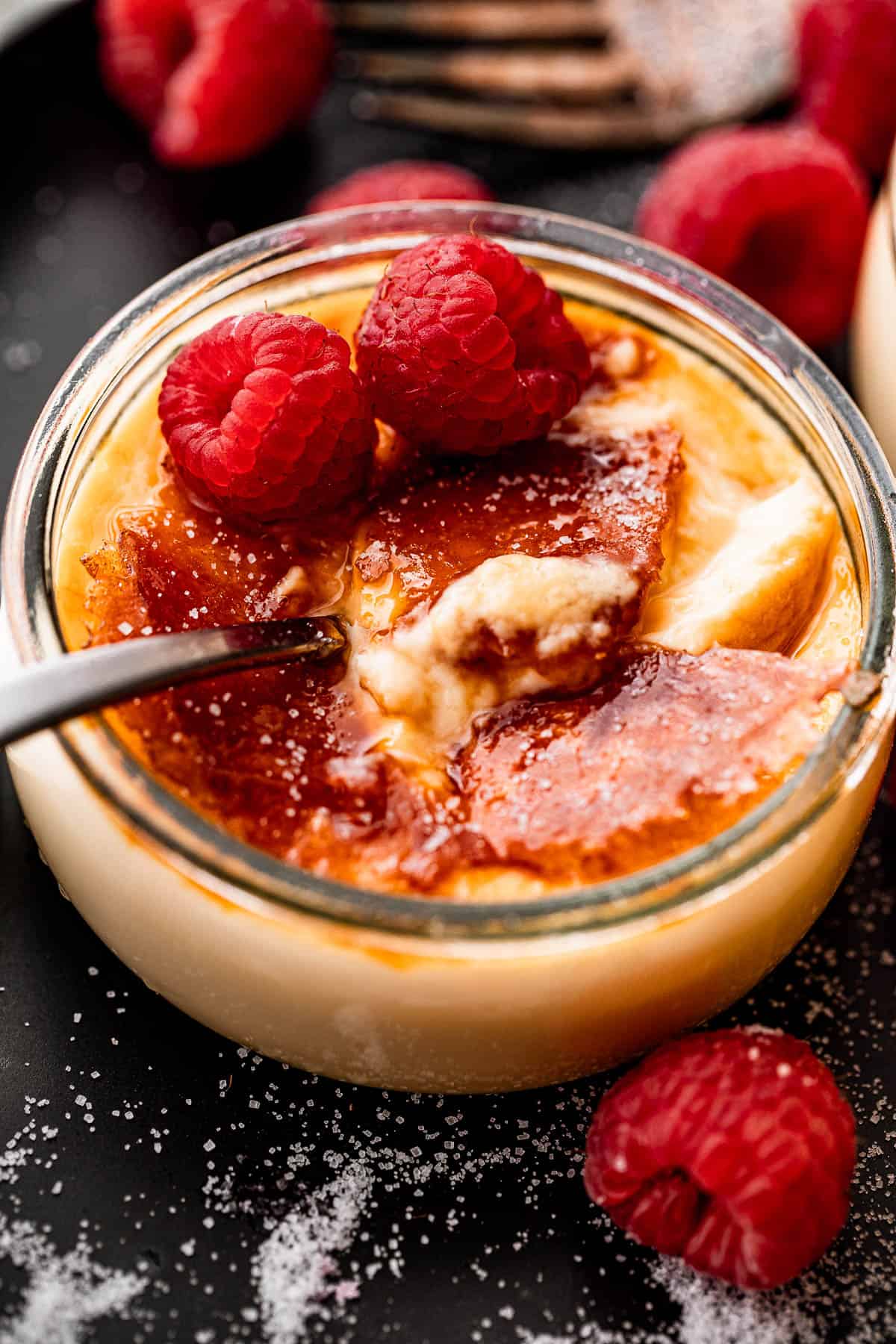 side shot of creme brulee in a glass jar, topped with two raspberries, and a spoon inside the jar.