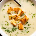 overhead close up shot of leek and potato soup served in a white bowl and garnished with croutons and chopped chives.