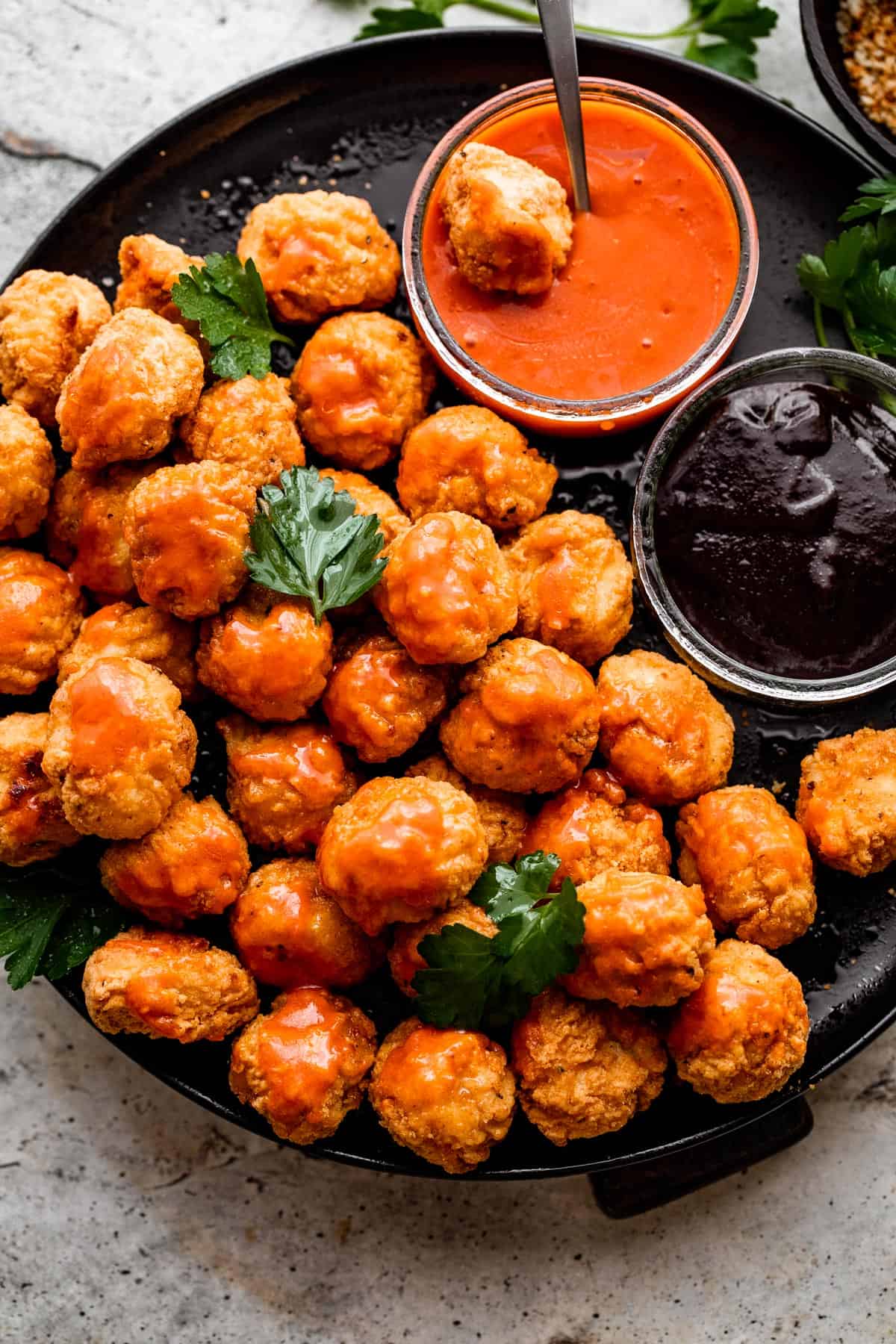 overhead shot of popcorn chicken arranged on a black plate with bowls of buffalo sauce and barbecue sauce placed right next to it.