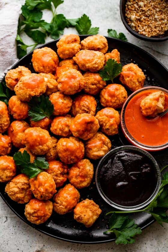 overhead shot of popcorn chicken arranged on a black plate with bowls of buffalo sauce and barbecue sauce placed right next to it.
