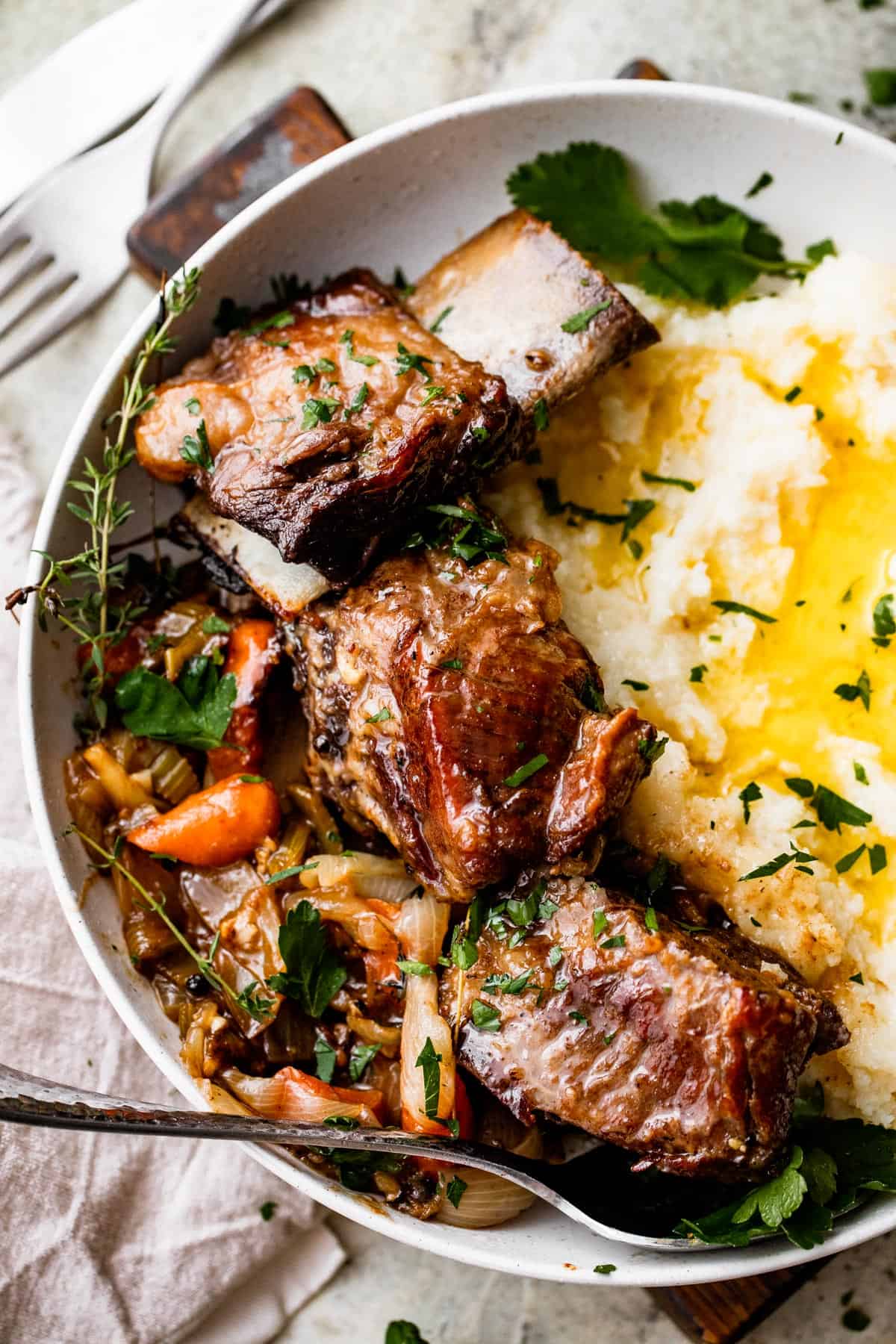 overhead shot of a dinner plate with three short ribs arranged around mashed potatoes, with veggies and onions set on the other side of the short ribs.