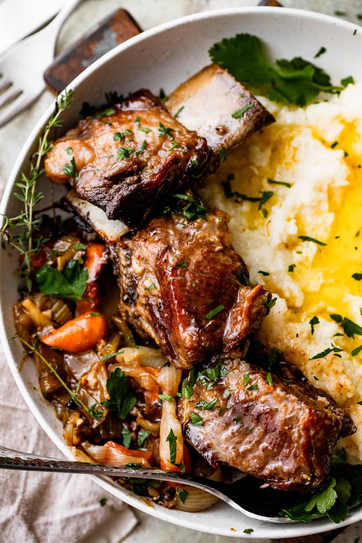 close up shot of a dinner plate with three short ribs arranged around mashed potatoes, with veggies and onions set on the other side of the short ribs.