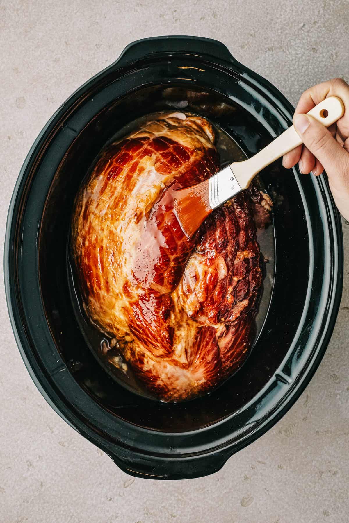 Overhead shot of a ham in a crockpot insert, being brushed with glaze.