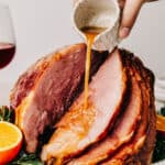 A woman's hand is pouring glaze from a small white measuring cup over a cooked spiral ham on a serving platter.
