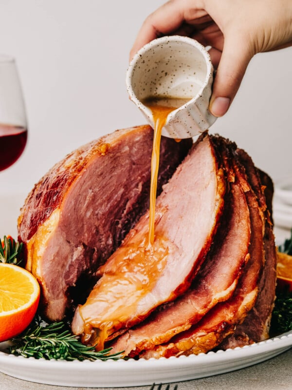 A woman's hand is pouring glaze from a small white measuring cup over a cooked spiral ham on a serving platter.