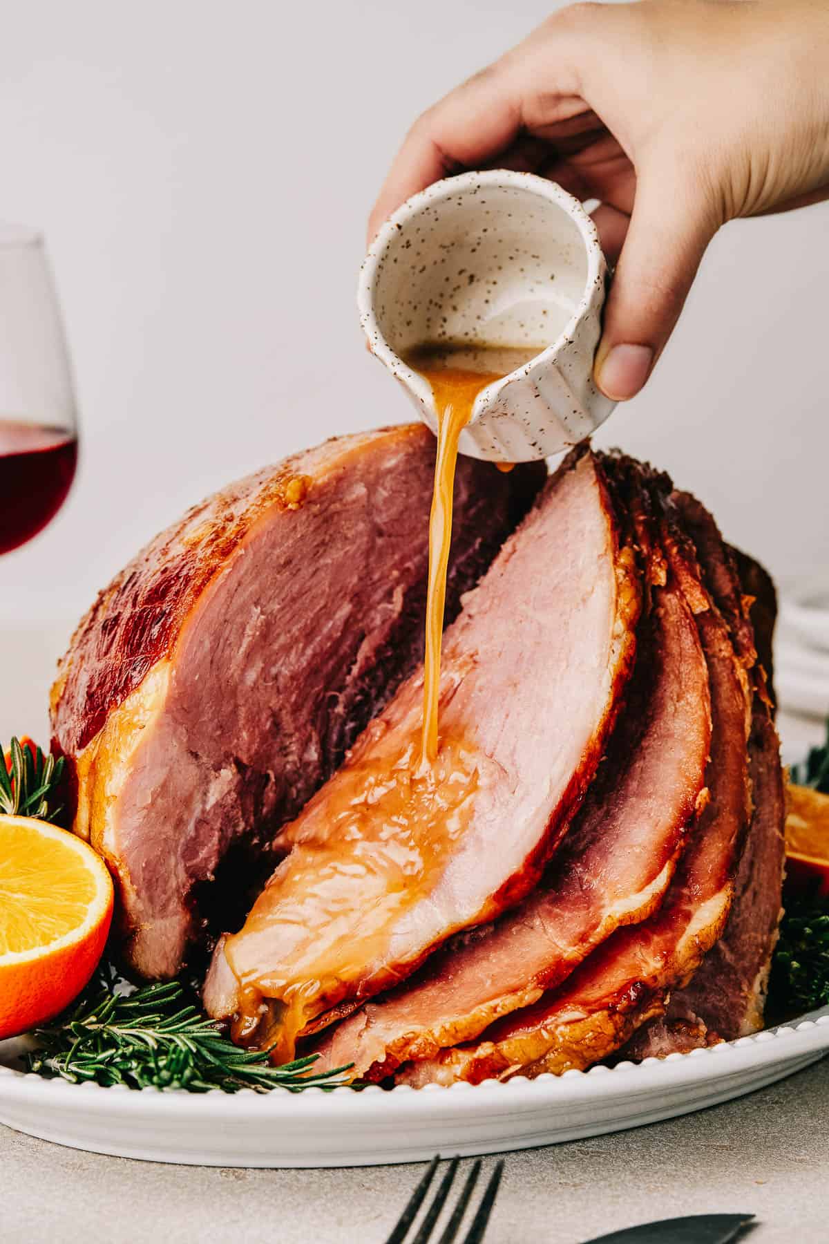 A woman's hand is pouring glaze from a small white measuring cup over a cooked spiral ham on a serving platter.