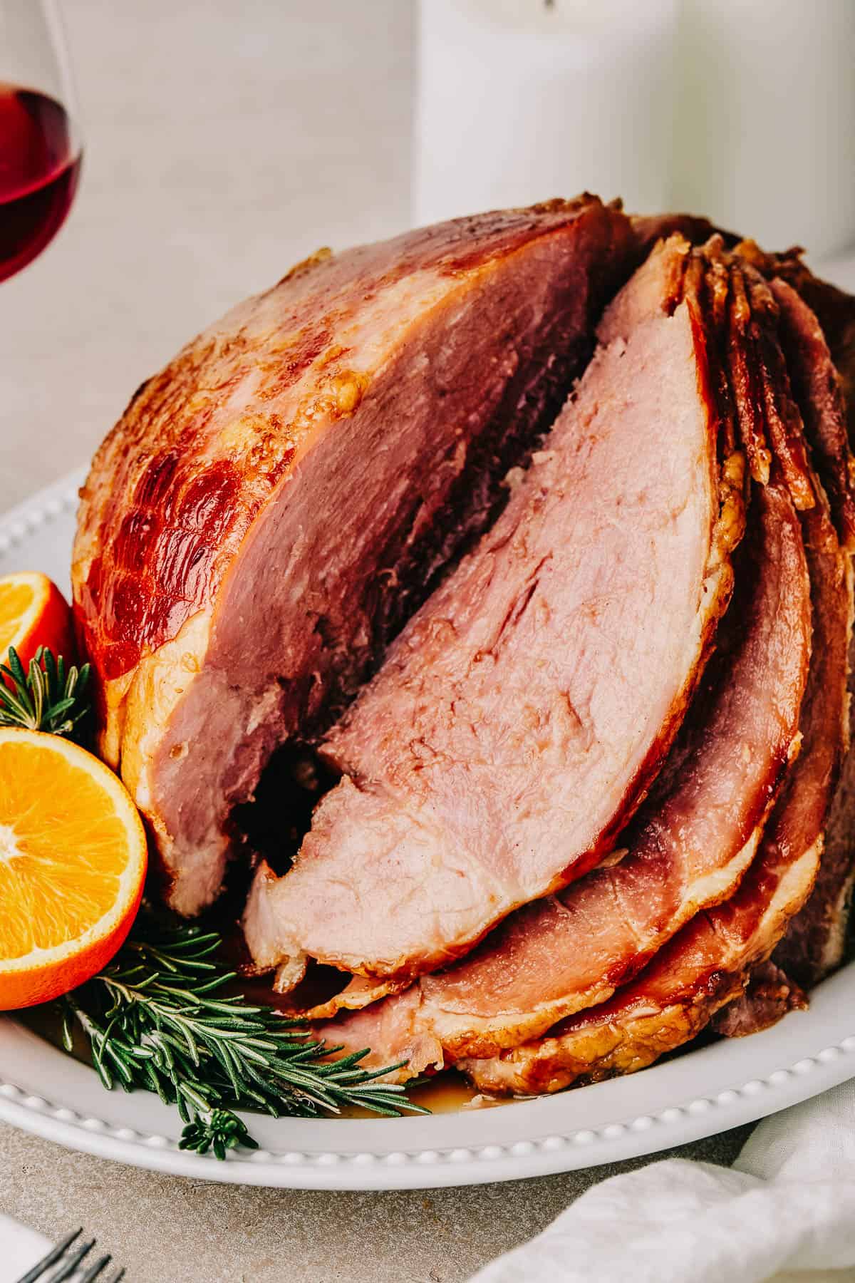 Sliced ham on a serving platter. A candle and a glass of wine are in the background of the shot, while a cup of glaze and several forks are in the foreground.