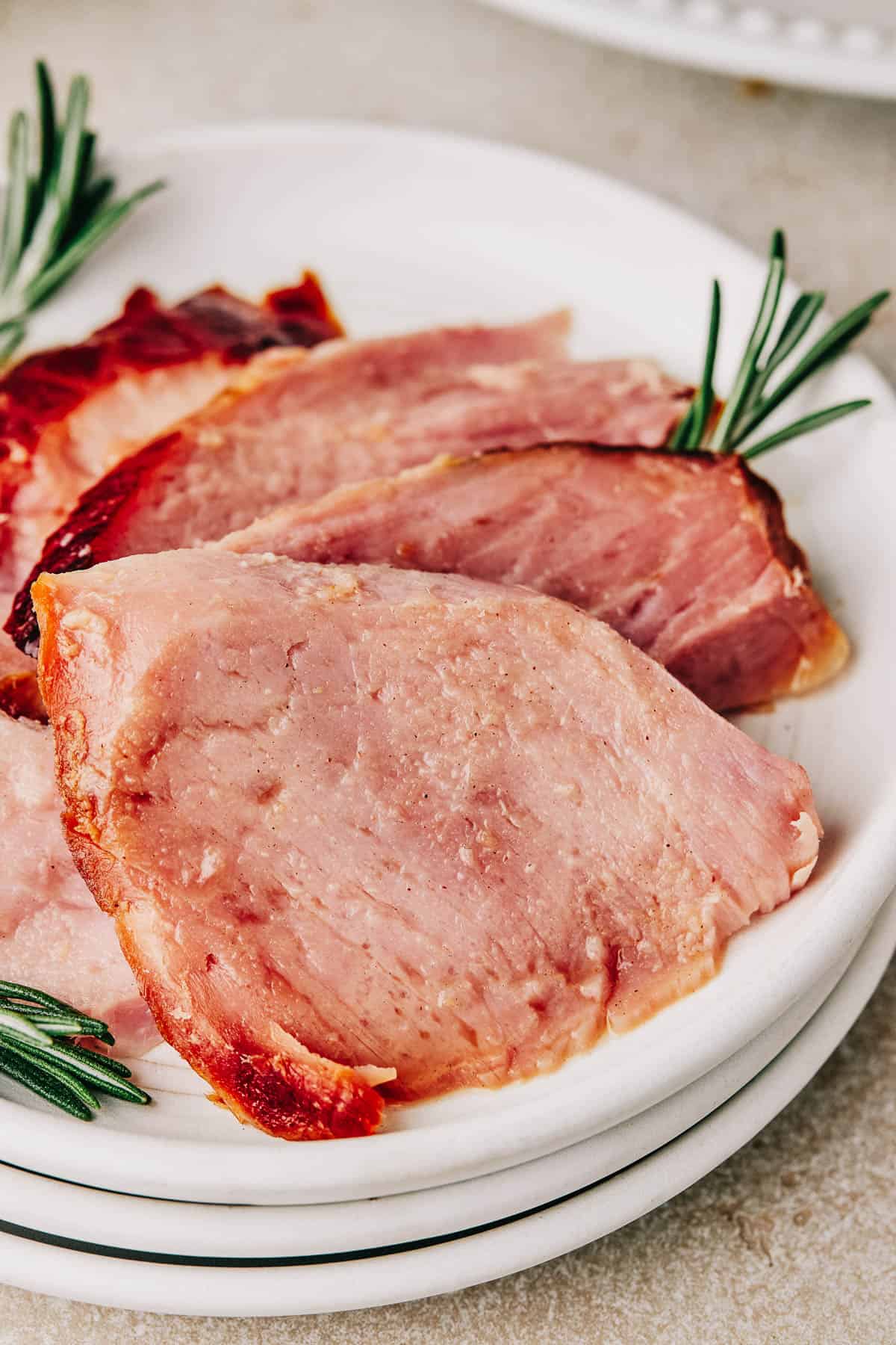 Four slices of ham on a small serving plate, garnished with rosemary. Forks, a cup of glaze, and the platter of ham are nearby on the table.