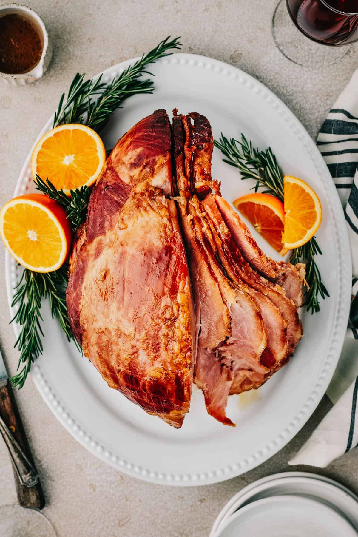 Overhead shot of a partially carved ham, with slices fanning out, on a serving platter garnished with orange slices and rosemary.