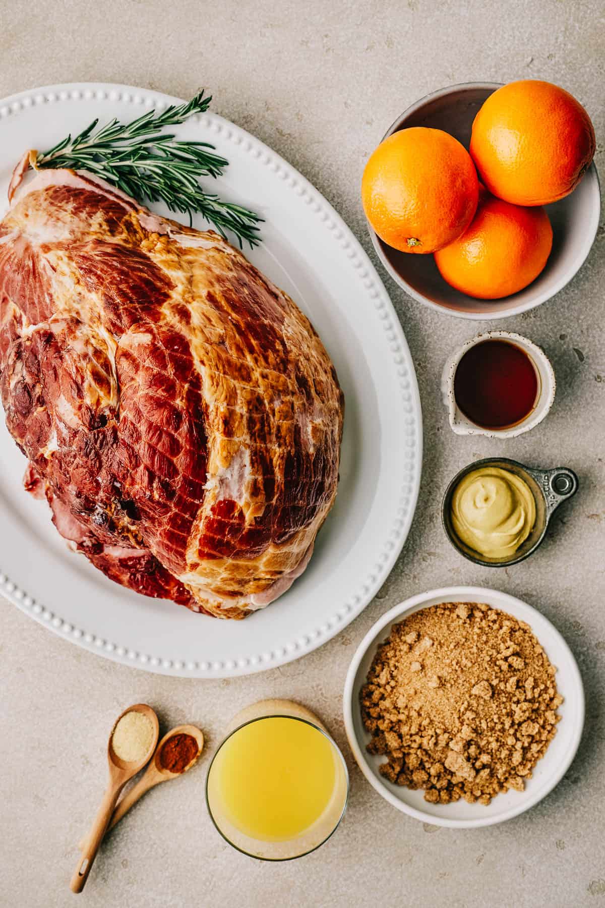 Clockwise from top left: A shank portion of ham, rosemary sprigs, oranges, maple syrup, dijon mustard, light brwon sugar, orange juice, cinnamon, garlic powder.