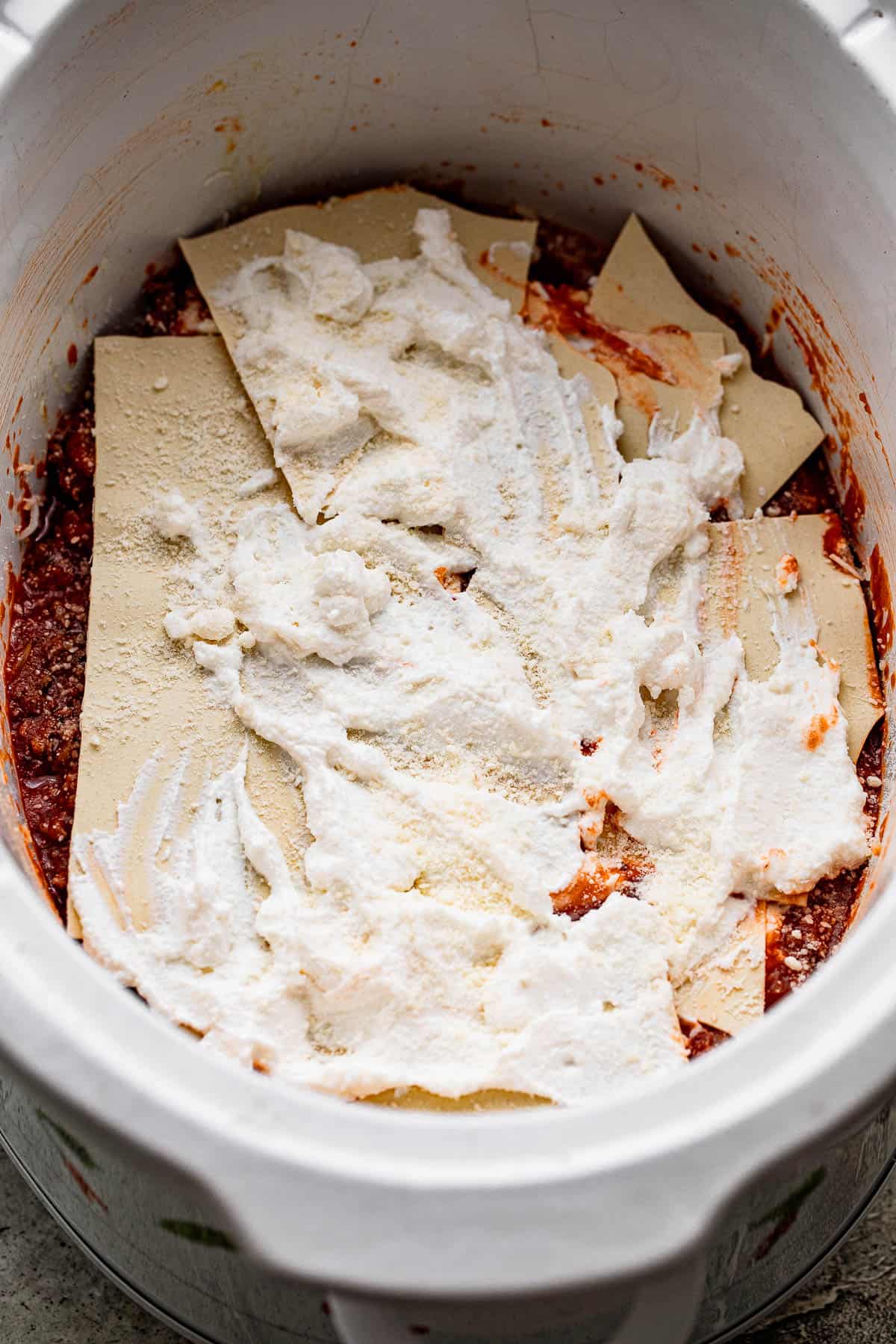 layers of tomato sauce, lasagna noodles, and ricotta cheese inside a slow cooker insert