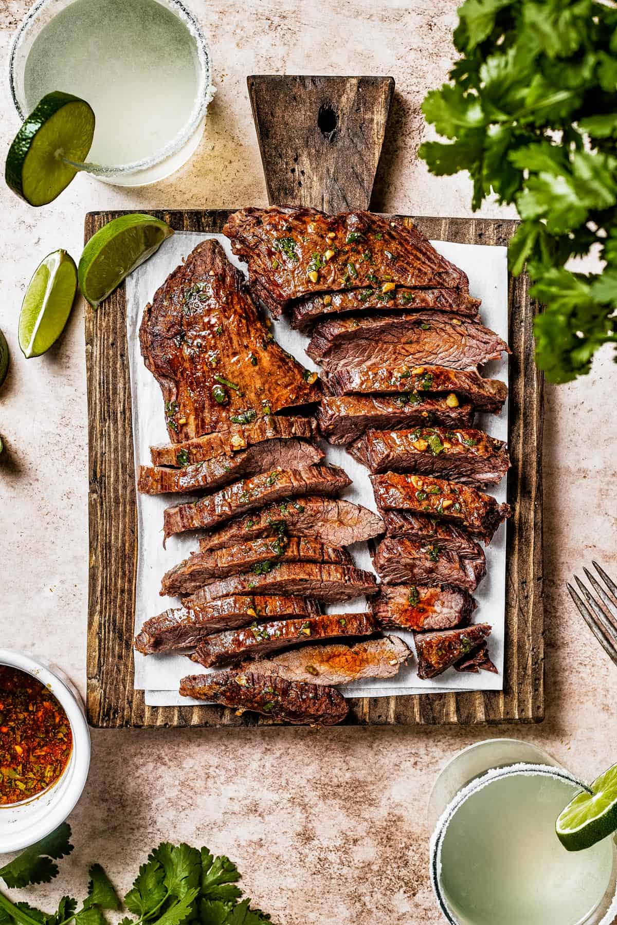 A cutting board with two large pieces of steak, sliced thinly. Limes an cilantro are on the work surface.