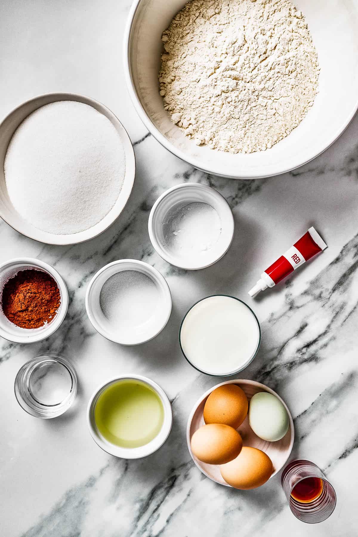 From top left: Sugar, baking soda, flour, cocoa powder, salt, milk, red food coloring, vinegar, oil, eggs, vanilla.
