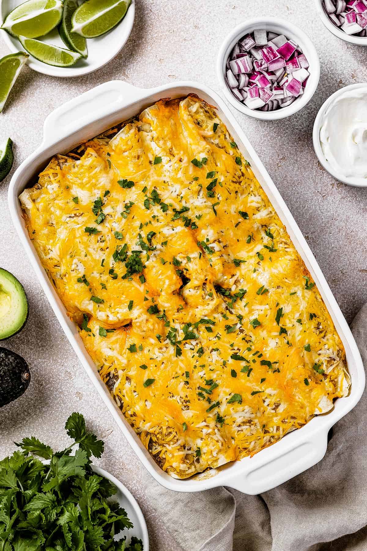 overhead shot of baked enchiladas in a white dish, with a small bowl of red onions set to the side of it, as well as a bowl with sour cream, and a bowl with lime wedges.