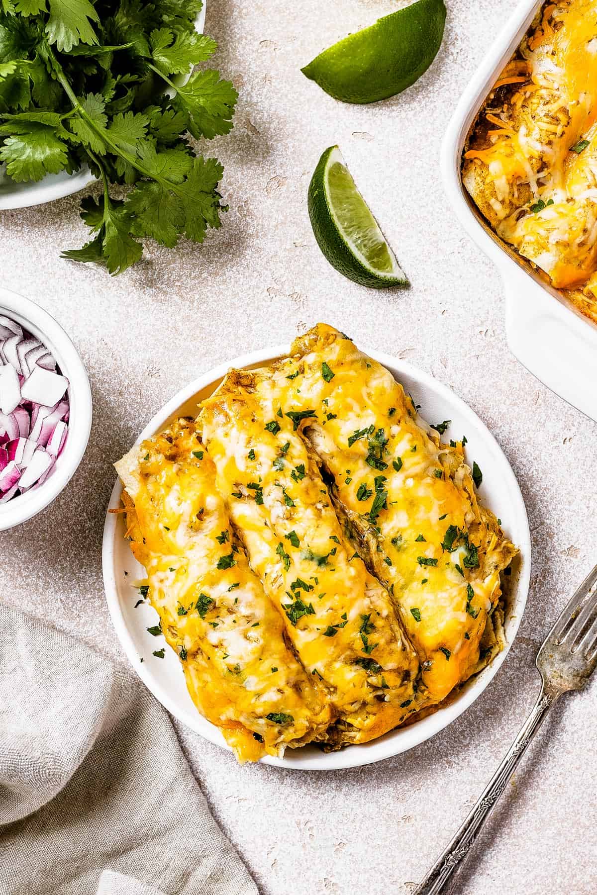A serving of chicken enchiladas on a small white plate, net to a dish of chopped onion and a dish of fresh cilantro.
